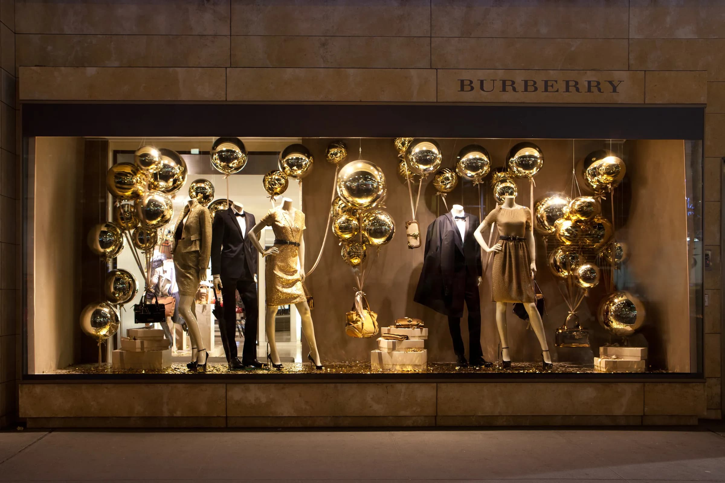 Burberry window at night filled with clusters of mirrored gold balloons around mannequins in evening wear